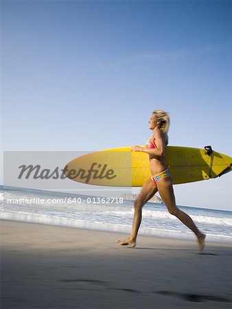 Profile of a mid adult woman running on the beach with a surfboard