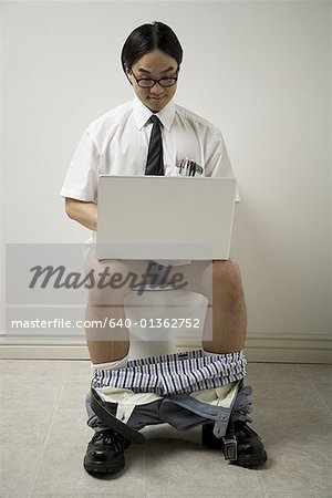 Close-up of a young man sitting on a toilet using a laptop