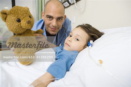 Girl lying in hospital bed, male intern next to her holding up teddy bear