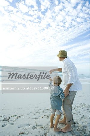 Senior man standing on beach with grandson, pointing toward distance