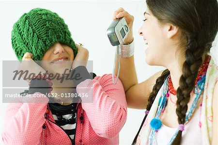 Two young female friends, one taking photo of the other while she pulls hat over eyes