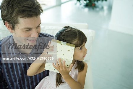 Girl holding up present to ear while father watches