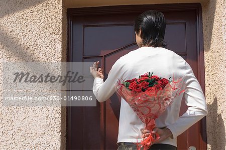 Rear view of a man hiding a bouquet of flowers behind his back and knocking the door