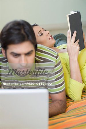 Mid adult man using a laptop with a young woman leaning on his back and reading a book
