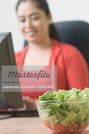 Close-up of a bowl of salad with a young woman using a computer in the background