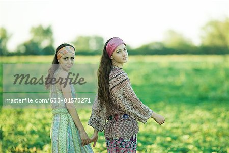 Young hippie women walking through field, holding hands, looking over shoulders at camera