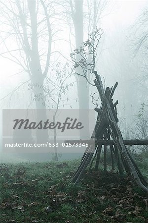 Misty landscape with teepee-shaped log structure in foreground
