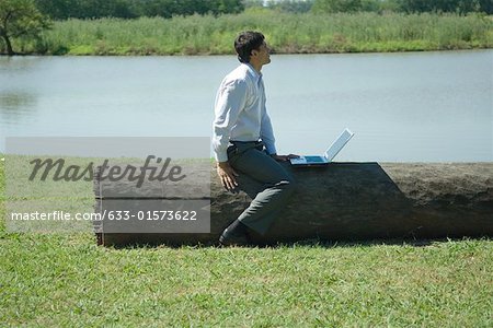 Businessman sitting on tree trunk with laZSop, looking away