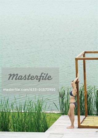 Young woman in swimsuit standing by edge of water, leaning against wooden structure