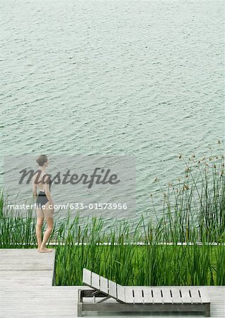Woman in swimsuit standing on deck, looking towards body of water