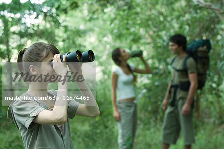Three hikers standing in forest, young woman in foreground looking through binoculars