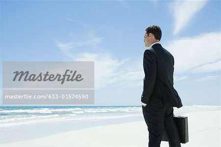 Businessman standing on beach, holding briefcase, facing ocean