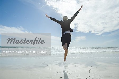 Businesswoman jumping in air on beach, rear view