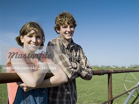 Brother and sister leaning on wooden fence in field with blue skies