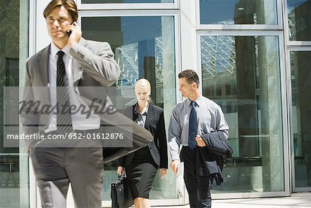 Male and female business colleagues talking as they leave office building, man using cell phone in foreground