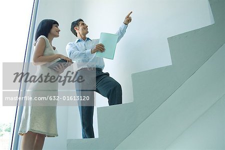 Businessman and young businesswoman standing on stairs, looking up, pointing