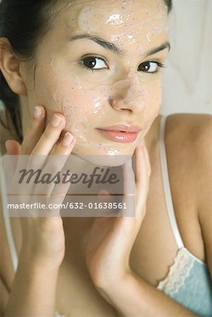 Young woman putting exfoliant mask on face, smiling at camera