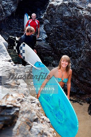 Three people carrying surfboards by large rocks smiling.