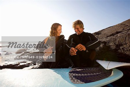 Couple sitting on beach with surfboard smiling.
