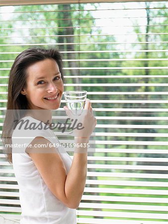 Woman standing by a window holding a glass of water smiling.