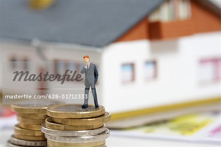 Businessman figurine on a stack of coins, miniature house in background