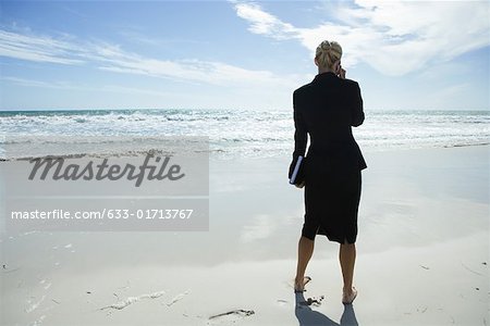 Femme d'affaires à l'aide de téléphone portable, se tenant debout sur la plage aux pieds nus, pleine longueur, vue arrière