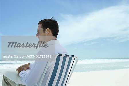 Mann sitzt im Klappstuhl am Strand, mit Stift und Papier, Seitenansicht
