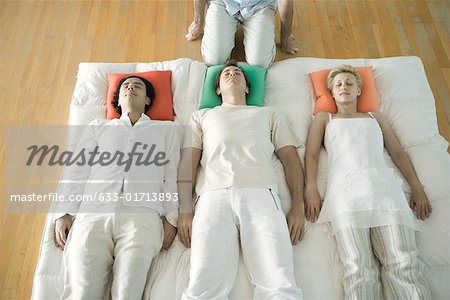 Alternative therapy session, three adults lying side by side while therapist kneels nearby, cropped