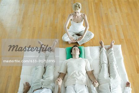 Alternative therapy session, three adults lying side by side, holding hands while therapist puts hands over man's head