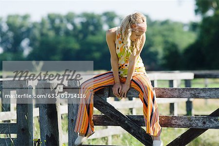 Young woman sitting on top of rural fence, looking down