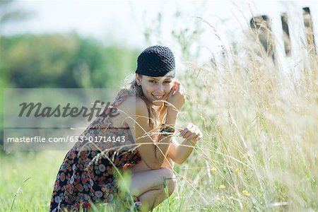 Jeune femme accroupie en champ, touchant les hautes herbes et souriant à la caméra