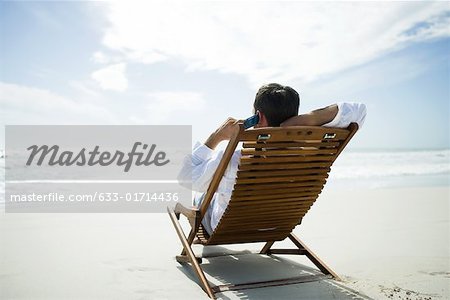 Mann sitzt im Stuhl am Strand, mit Handy, Rückansicht