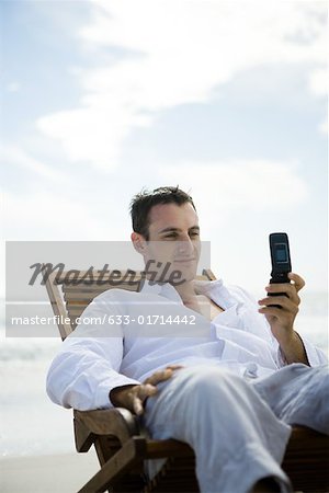 Junger Mann sitzt in einem Stuhl am Strand, mit Handy