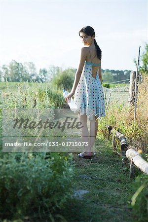 Young woman standing in garden with bottle of water, looking over shoulder at camera