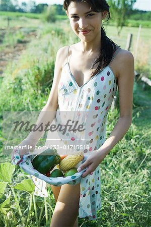 Young woman standing in garden, holding fresh produce in dress, smiling at camera
