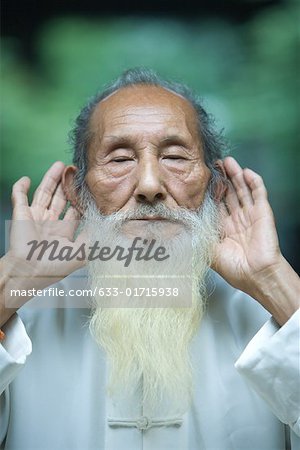 Elderly man in traditional Chinese clothing holding hands behind ears