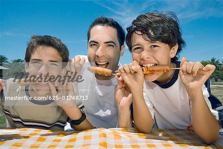 Portrait d'un homme adult moyen avec ses deux fils, manger des saucisses dans un pique-nique