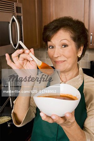 Portrait d'une femme senior tenant un bol de soupe de tomate et une cuillère de bois dans la cuisine