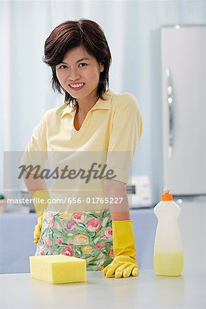 Woman in kitchen, wearing gloves, looking at camera, cleaning detergent and sponge on kitchen counter