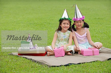 Two girls wearing party hats sitting on picnic blanket, surrounded by gifts