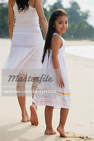 Mother and daughter walking down beach, daughter looking back towards camera smiling