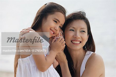 Mother and daughter on beach with ears up to a conk shell, listening to the ocean