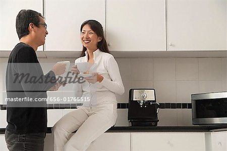 Femme assise sur le comptoir dans la cuisine avec l'homme debout à côté d'elle, regardant les uns les autres, tenant des tasses à café et souriant