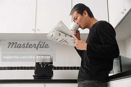 Man in kitchen, drinking coffee and reading paper