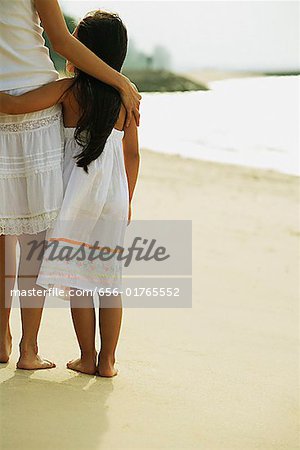 Mother and daughter standing on beach, arms around each other, rear view