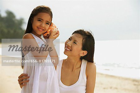 Mère et fille sur la plage, fille avec coque blanche spongieuse à l'oreille à l'écoute de l'océan