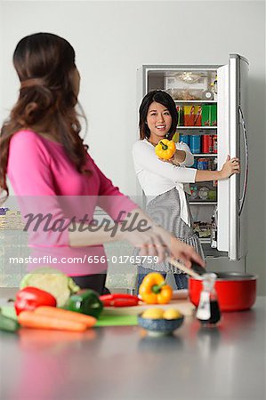 Mother and adult daughter in kitchen, daughter at refrigerator holding bell pepper