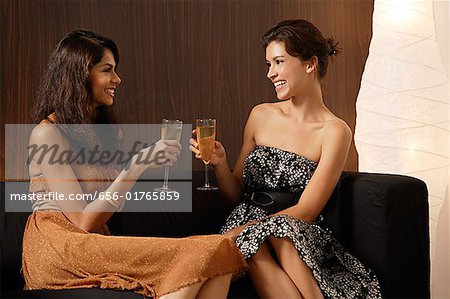 Two girls sitting on couch, toasting each other with champagne, smiling at each other