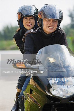 Man and woman riding motorcycle, wearing helmets, looking at camera
