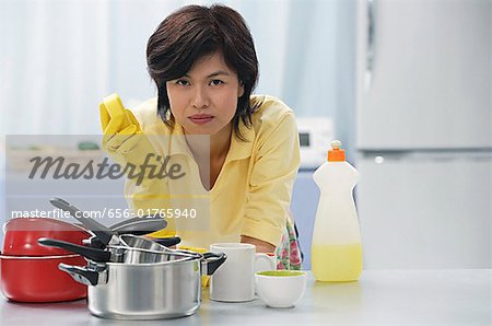 Woman leaning on kitchen counter, holding cleaning sponge, stack of pots and pans in front of her
